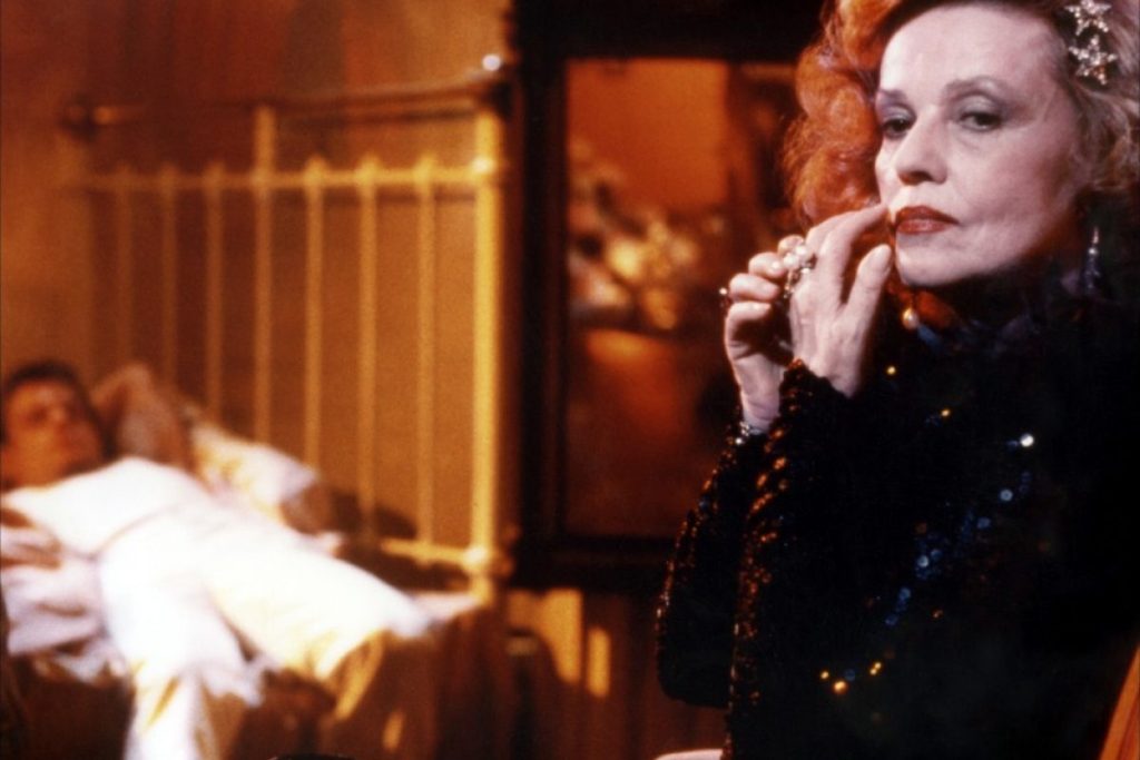 Scène de Querelle de Brest : Le heros se repose au second plan allongé sur un lit en métal blanc, les bras sous la tête, à droite de l'image, au premier plan, une femme d'âge mûr maquillée regarde pensivement devant elle.